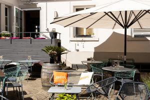 Sonniger Terrassenbereich mit einer Metallbank samt Aperol-Kissen im Vordergrund. Dahinter führt eine kleine Treppe zu einer geöffneten Tür, rechts spendet ein großer Sonnenschirm Schatten.