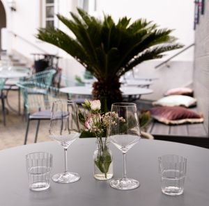 Nahaufnahme von leeren Gläsern und einer Blumenvase auf einem grauen Tisch, im unscharfen Hintergrund sind eine Palme und weitere Sitzgelegenheiten zu sehen.