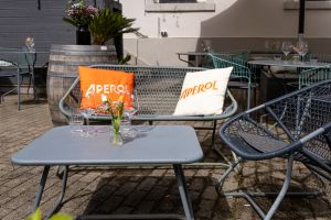 Gemütliche Sitzecke im Freien mit einer grauen Metallbank, verziert mit auffälligen, orangefarbenen und weißen Aperol-Kissen, davor ein niedriger grauter Beistelltisch.