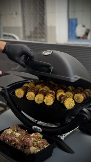 Maißkolben auf dem Grill, davor rohe Grillspieße in einer Schale